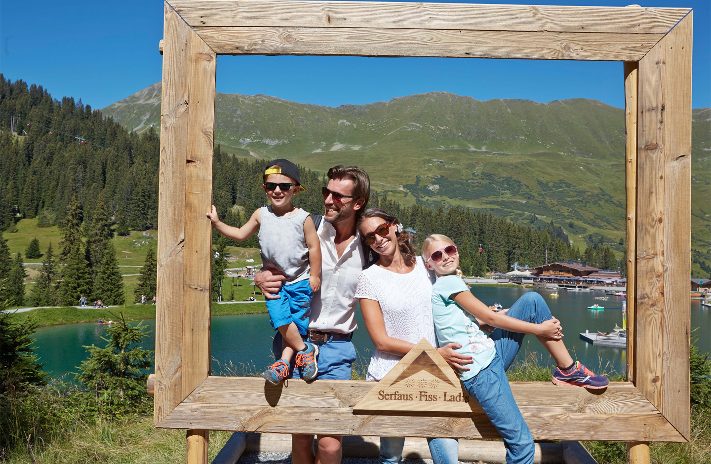 Eine Familie posiert in einem großen Holzrahmen vor einer Berg- und Seenlandschaft in Serfaus-Fiss-Ladis. Im Hintergrund sind grüne Almwiesen, Wälder, Berge und ein See mit Ausflugsgästen zu sehen.